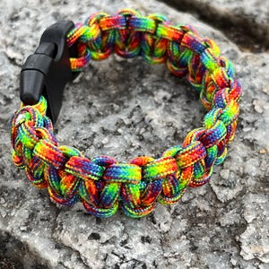 Op de afbeelding: Een regenboogkleurige paracordarmband met een zwarte plastic sluiting. De armband is gemaakt van strak geweven nylon koord in verschillende kleuren, waaronder rood, oranje, geel, groen, blauw en paars. De armband wordt getoond op een grijze, getextureerde ondergrond.