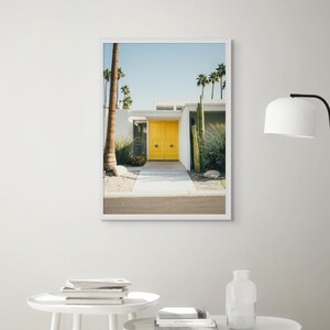 Peut inclure: Une photographie encadrée d'une porte jaune avec un cadre blanc, placée contre un mur blanc. La porte est sur une maison avec un extérieur blanc et un palmier au premier plan. La maison est entourée d'un paysage désertique.