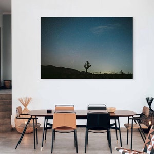 Peut inclure: Une salle à manger avec une table et des chaises en bois, un tapis et un tabouret en osier. Une grande impression d'un paysage désertique avec un arbre solitaire sous un ciel étoilé est accrochée au mur.