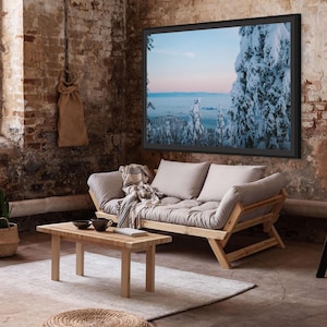 May include: A light brown futon sofa with a wooden frame and a light grey cushion. The sofa is in a room with a brick wall, a large framed picture of a snowy forest, and a black ladder.