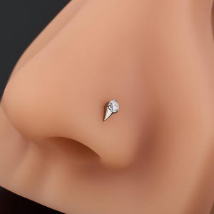 Può includere: Un piercing al naso color argento con una piccola pietra preziosa rotonda e trasparente incastonata in una base metallica conica a punta. Il gioiello è esposto su un manichino beige a forma di naso. Il testo OUFER BODY JEWELRY è visibile.