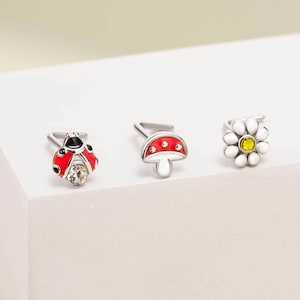 Op de afbeelding: Drie zilverkleurige oorclips met op de natuur geïnspireerde ontwerpen. Eén is een rode lieveheersbeestje met zwarte stippen en een heldere edelsteen. Een andere is een rode paddenstoel met witte stippen. De derde is een witte madelief met een gele kern.