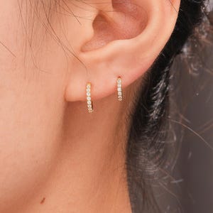 May include: A close-up of a person's ear adorned with two small, gold hoop earrings. Each earring is encrusted with small, sparkling white gemstones. The earrings are a simple, elegant design, perfect for everyday wear. The jewelry is from OUFER BODY JEWELRY.