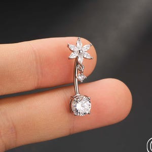 May include: A silver belly button ring with a flower design at the top and a dangling gem. The bottom features a large, round, clear gem. The jewelry is held by a person's fingers. The brand name "OUFER BODY JEWELRY" is visible.