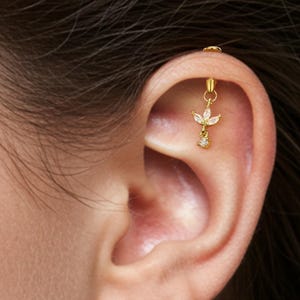 Op de afbeelding: Een goudkleurige helix-oorbel met een bloemmotief en een bungelende heldere edelsteen. De oorbel is aan het oor bevestigd. De merknaam "OUFER BODY JEWELRY" is rechtsonder zichtbaar.