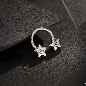 May include: A silver-colored horseshoe-shaped body jewelry piece with star-shaped ends. Each star features a spiral design. The jewelry is displayed on a dark, textured surface. The brand name "OUFER BODY JEWELRY" is visible in the bottom right corner.