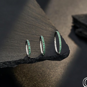 Puede incluir: Tres pendientes de aro plateados, cada uno adornado con una fila de pequeñas piedras preciosas verdes brillantes. Los pendientes se exhiben sobre una superficie oscura y texturizada. El logotipo "OUFER BODY JEWELRY" es visible.