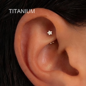 Può includere: Primo piano di un orecchio con un piccolo orecchino in titanio a forma di stella. La parola "TITANIUM" è stampata in bianco in alto a sinistra. L'orecchio ha un piccolo orecchino a cerchio dorato nell'elica. Il logo del marchio "OUFER BODY JEWELRY" è in basso a destra.