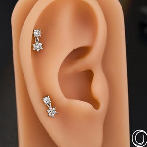 May include: Two silver-coloured flower-shaped earrings with clear gemstones are displayed on a tan ear model. The earrings have a stud design with a dangling flower. The background is black.