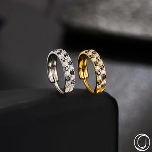 May include: Two hoop earrings, one silver and one gold, each adorned with a double row of black and clear gemstones. The earrings are displayed on a black surface, with the brand name "OUFER BODY JEWELRY" visible.
