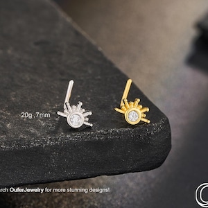 Może przedstawiać: Dwa kolczyki do nosa, jeden srebrny i jeden złoty, każdy z motywem promieni słonecznych i centralnym, przezroczystym kamieniem. Srebrny kolczyk jest oznaczony "20g, 7mm". Obraz zawiera również tekst "Search OuferJewelry for more stunning designs!"