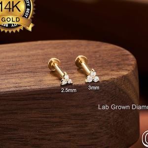 May include: Two gold-coloured labret studs, each with a triangular cluster of small, clear gemstones. One stud is 2.5mm, the other is 3mm. The image includes a gold seal that reads "14K GOLD" and the text "Lab Grown Diamond".