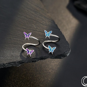 May include: Two silver-toned body jewelry pieces, each featuring a spiral design with two butterfly accents. One piece has purple butterflies, the other has blue butterflies. The jewelry is displayed on a dark, textured surface.