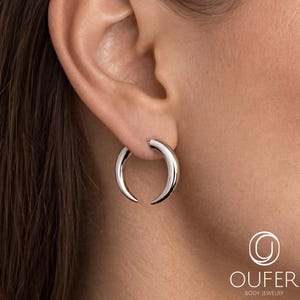 May include: A polished silver-toned crescent moon hoop earring with a smooth, curved design that tapers to pointed ends. The earring is worn in the earlobe, featuring a sleek, metallic finish and a minimalist, open-hoop structure.