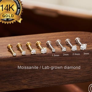 Puede incluir: Una colección de labrets dorados y plateados con piedras de moissanita transparentes, que varían de 1,5 mm a 3 mm. Los pendientes se exhiben sobre una superficie de madera oscura, con un sello dorado que dice "14K Gold". También se ve el texto "Moissanite / Lab-grown diamond".