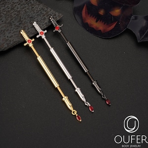 Puede incluir: Tres joyas para el cuerpo con forma de espada en oro, plata y negro, cada una con una gema roja. Las joyas se exhiben sobre una superficie negra con decoraciones temáticas de Halloween, incluyendo un murciélago y una calabaza.