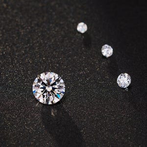 May include: Four sparkling, round-cut diamonds of varying sizes are arranged on a dark, textured surface. The largest diamond is in the foreground, with three smaller diamonds trailing behind it, all reflecting light.