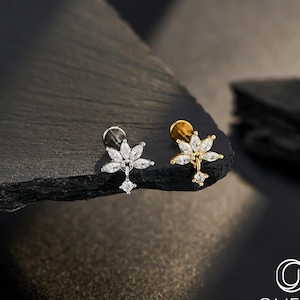 May include: Two stud earrings, one silver and one gold, each with a floral design and clear gemstones. The earrings are presented on a dark, textured surface, highlighting their intricate details and elegant style. The earrings are body jewellery.