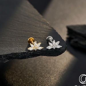 May include: Two pieces of body jewelry. One is gold-toned, the other silver-toned. Each features a floral design with clear, sparkling stones. The jewelry is displayed on a dark, textured surface, with the brand name "OUFER BODY JEWELRY" visible in the lower right corner.