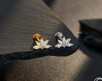 Puede incluir: Dos piezas de joyería corporal. Una es dorada, la otra plateada. Cada una presenta un diseño floral con piedras claras y brillantes. La joyería se exhibe sobre una superficie oscura y texturizada, con la marca "OUFER BODY JEWELRY" visible.