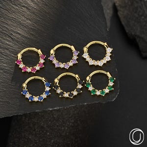 May include: Six gold-tone hoop rings, each featuring a segment of five small, round-cut gemstones. The stones are arranged in different colors: pink, light purple, clear white, blue, black, and green. Each hoop has a hinged closure mechanism.