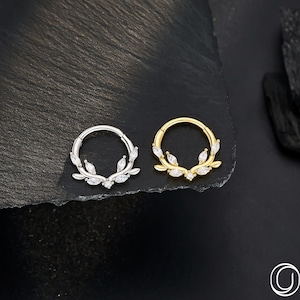 Op de afbeelding: Twee lichaamsjuwelen ringen, &eacute;&eacute;n zilver en &eacute;&eacute;n goud, elk met een bladvormgeving en heldere edelstenen. De ringen staan tegen een donkere, getextureerde achtergrond, met het merklogo rechtsonder.