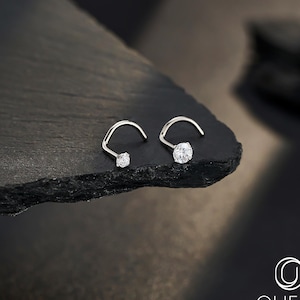 May include: A pair of silver nose studs, each featuring a curved design with a sparkling, clear gemstone. One stud has a smaller stone, while the other has a larger one. The jewelry is displayed on a dark, textured surface.