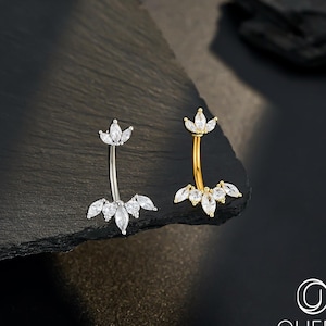 May include: Two belly button rings, one silver and one gold, each featuring clear, marquise-cut gemstones in a floral pattern. The jewellery is presented on a dark, textured surface. The brand logo is visible in the bottom right corner.