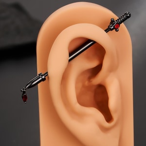 Puede incluir: Pendiente industrial negro con extremos en forma de espada y detalles rojos. El pendiente se muestra en un modelo de oreja bronceado, mostrando el diseño y los detalles de la joya. Una pieza de joyería corporal única.