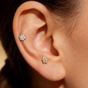 May include: Close-up of an ear adorned with two flower-shaped stud earrings. One earring is set with clear gemstones, while the other is gold-toned with similar gemstone accents. The earrings are delicate and add a touch of sparkle.