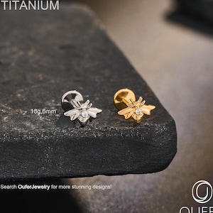 Puede incluir: Dos pendientes para el cuerpo con forma de abeja, uno plateado y otro dorado, cada uno con una pequeña gema transparente. La palabra "TITANIUM" está encima de los pendientes. El texto "18g,6mm" está junto al pendiente plateado. El texto "Search OuferJewelry for more stunning designs!" está debajo.