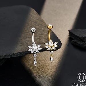 Pode incluir: Dois piercings no umbigo. Um é prateado, o outro dourado. Cada um apresenta um design floral com cristais claros e brilhantes. O anel prateado tem um cristal pendurado, enquanto o anel dourado tem um cristal pendurado. O logotipo "OUFER BODY JEWELRY" está no canto inferior direito.