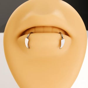 May include: A silver-toned horseshoe-shaped body jewelry piece featuring two pointed, fang-like ends. The jewelry is displayed on a smooth, light-toned silicone model of a mouth, highlighting the curved metal bar and the sharp, tapered tips.