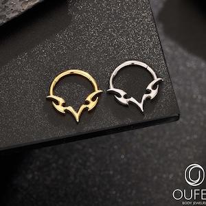 Op de afbeelding: Twee septumringen met een vlamontwerp. Eén ring is goudkleurig, de andere zilverkleurig. De ringen worden getoond op een donker, getextureerd oppervlak. De ringen zijn lichaamsjuwelen.