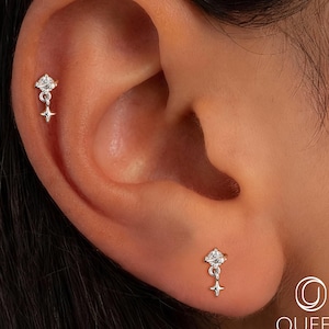 May include: A close-up of an ear with two silver earrings. Each earring features a small, clear gemstone set in a starburst design, with a dangling star charm. The earrings are delicate and elegant.