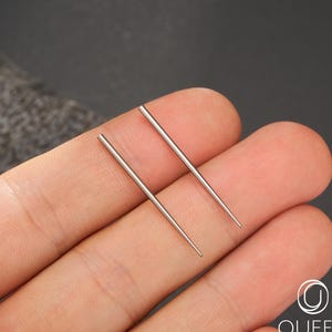 May include: Two silver-colored body jewelry needles are held in a hand. The needles are straight and have a pointed tip. The needles are thin and appear to be made of metal. The background is a dark gray color. The logo "OUFER BODY JEWELRY" is in the bottom right corner.