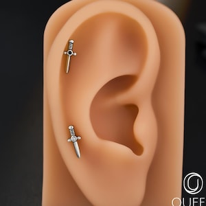 May include: Two silver dagger-shaped earrings displayed on a tan ear model. One earring has a black gem, the other a clear gem. The earrings are body jewellery. The background is black.