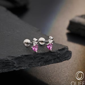 May include: A pair of silver-toned earrings with a heart-shaped pink gemstone and a small clear stone. The earrings have a flat back and a post for piercing. The earrings are displayed on a dark, textured surface.