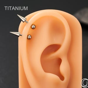 Puede incluir: Joyas para el cuerpo de titanio, que incluyen dos pendientes de punta plateados y dos pendientes de bola pequeños, se muestran en un modelo de oreja bronceado. La palabra "TITANIUM" está impresa en blanco en la parte superior. Las joyas son de OUFER BODY JEWELRY.