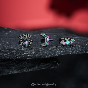 Puede incluir: Tres piezas de joyería corporal negra con un acabado brillante. Las piezas son una araña, una luna creciente y un murciélago. La purpurina es una mezcla de verde, azul y rosa.