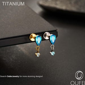 Puede incluir: Dos pendientes de titanio con piedras preciosas en forma de lágrima de color azul claro. Cada pendiente tiene un pequeño dije colgante con una piedra preciosa más pequeña. La palabra "TITANIUM" está en la parte superior. El texto "Search OuferJewelry for more stunning designs!" está en la parte inferior.