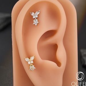 May include: Two gold-coloured butterfly and flower-shaped ear piercings with clear gemstones are displayed on a tan ear model. The piercings feature a butterfly design above a flower. The OUFER Body Jewelry logo is visible in the lower right corner.