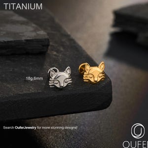 Puede incluir: Dos pendientes con forma de zorro, uno plateado y otro dorado, se muestran sobre una superficie oscura. El texto "TITANIUM" está en la parte superior. La talla es 18g, 6mm. El logotipo de Oufer Body Jewelry está en la esquina inferior derecha.