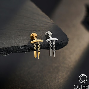 May include: A pair of stud earrings, one gold and one silver, each with a horizontal bar design, chains, and cone pendants. They are presented on a dark, textured surface. The brand logo is visible in the bottom right corner of the image.