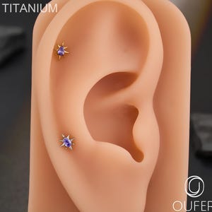 Peut inclure: Deux clous d'oreilles en forme d'étoile avec des centres violets sont présentés sur une oreille prothétique. Les clous sont de couleur or et ont plusieurs pointes. Le mot "TITANIUM" est visible dans le coin supérieur gauche. La marque "OUFER BODY JEWELRY" est dans le coin inférieur droit.