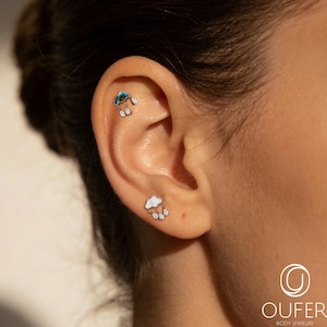 May include: Two silver-toned stud earrings shaped like clouds with dangling teardrop accents. The upper earring features a blue iridescent inlay, while the lower earring has a white pearlescent inlay. Both pieces include small clear stones.
