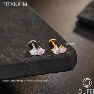 Op de afbeelding: Twee titanium body jewelry studs. Eén is zilverkleurig, de andere goudkleurig. Elk heeft een helder en roze hartvormig edelsteenontwerp. De tekst "TITANIUM" staat bovenaan. De tekst "18g,6mm" is zichtbaar. De tekst "Search OuferJewelry for more stunning designs!" staat onderaan.