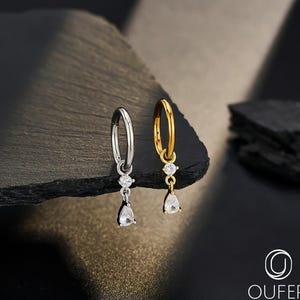 Op de afbeelding: Twee oorringen, één zilver en één goud, elk met een druppelvormige, heldere edelsteen. De oorbellen worden gepresenteerd op een donker, getextureerd oppervlak tegen een zwarte achtergrond. De gouden oorring heeft een bijpassende gouden ketting.