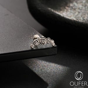May include: A silver body jewellery item with a series of tiny rose motifs. The jewellery is positioned on a dark, reflective surface, against a blurred black backdrop. The brand logo "OUFER BODY JEWELRY" is visible in the bottom right corner.