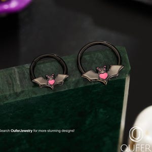 以下が含まれることがあります： ピンクのハートのデザインが施された、黒いコウモリ型のボディジュエリーリングが2つ。リングは濃い緑色の表面に表示されています。「Search OuferJewelry for more stunning designs!」というテキストが表示されています。